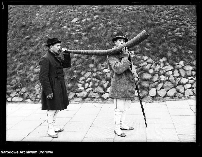 Archiwalna fotografia: dwaj mężczyźni w kapeluszach i długich płaszczach grają na bardzo długich drewnianych trąbach opartych o ziemię