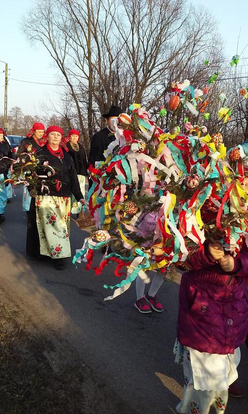 Fotografia: pochód ludzi niosących wysokie gałęzie obwieszone kolorowymi wstążkami i kukłami